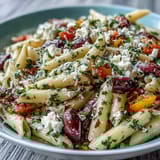 Vibrant Greek pasta salad with olives and feta, colorful veggies, and tangy dressing in a white bowl.