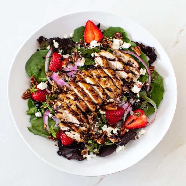 Sliced grilled chicken over mixed greens with strawberries, goat cheese, and a honey balsamic glaze on a plate.